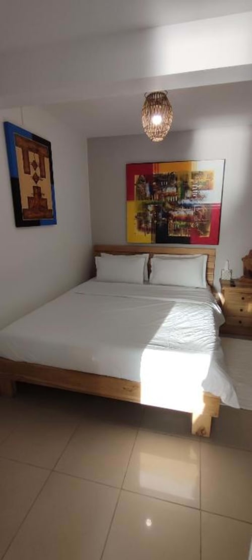 Chambre lumineuse avec lit en bois, draps blancs, œuvres d'art colorées, suspension et carrelage à Clémara Evasion