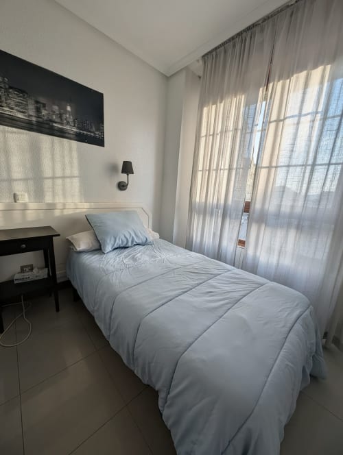 Habitación individual luminosa en Alojamiento La Ballena en Santoña con ropa de cama azul, escritorio y vistas panorámicas a través de cortinas translúcidas