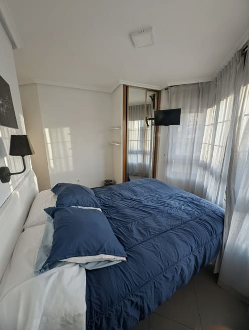 Moderno dormitorio con cama doble, ropa de cama azul, TV y grandes ventanales en Alojamiento La Ballena de Santoña