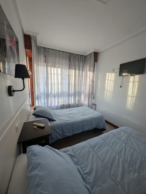 Dormitorio doble en Alojamiento La Ballena de Santoña con dos camas, lámpara de pared, TV y ventana con cortinas translúcidas