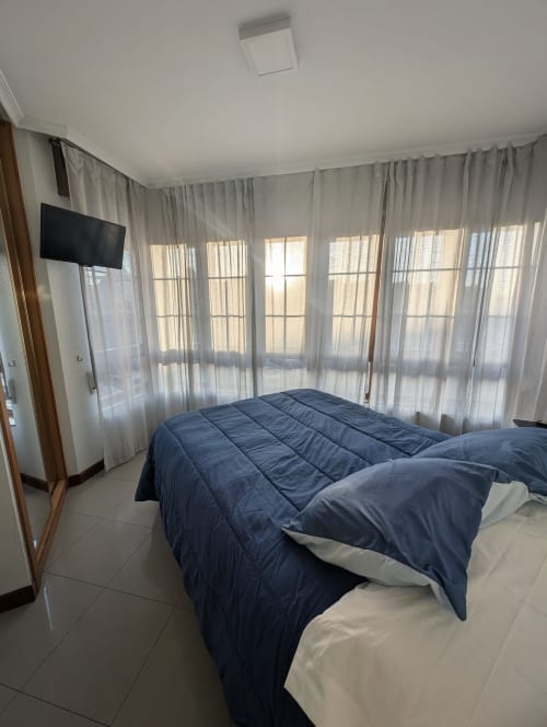 Luminosa habitación doble con ropa de cama azul, vistas a la ciudad, cortinas traslúcidas, TV y suelo de baldosas en Alojamiento La Ballena