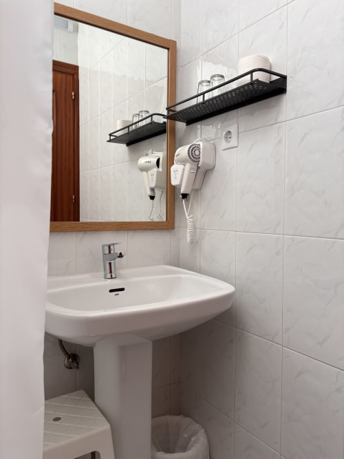 Baño moderno del Hostal Las Dunas con lavamanos pedestal, espejo, estantes negros y secadores de pelo montados en la pared