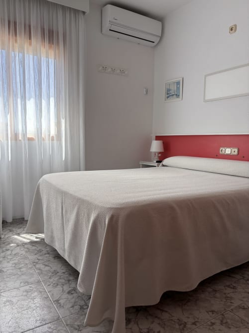 Habitación moderna en Hostal Las Dunas con cabecero rojo, ropa de cama blanca, aire acondicionado y luz natural de las cortinas