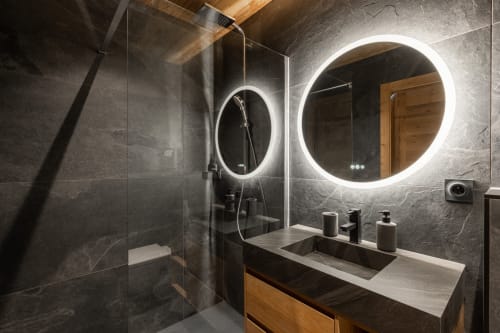 Modern bathroom in Altitudia villa with dual illuminated mirrors, dark marble tiles, wooden vanity, and rainfall shower at