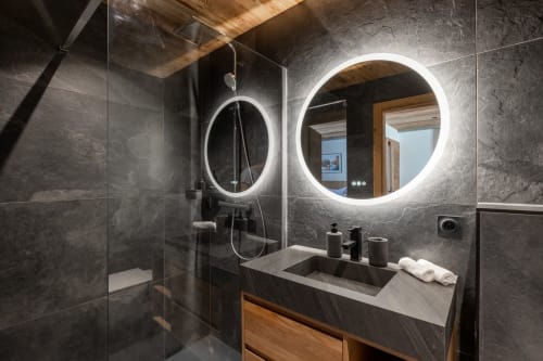 Modern luxury bathroom in Altitudia chalet featuring LED-lit round mirrors, dark slate tiles, and wooden vanity with stone