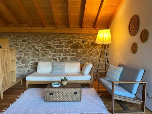 Aconchegante sala de estar na Quinta da Mata de Baixo com paredes de pedra, vigas de madeira, sofá creme e almofadas listradas com vista para o