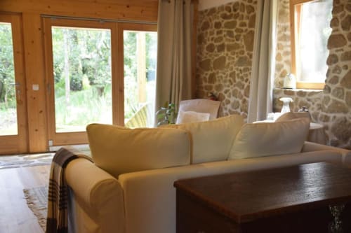 Sala acolhedora com sofá creme, lareira em pedra e portas de madeira com vista para o jardim da Quinta da Mata de Baixo