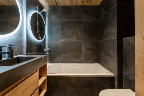 Modern bathroom in Chalet Magy at Altitudia with marble tiles, illuminated round mirror, wooden vanity, and white bathtub