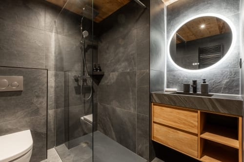 Modern luxury bathroom with dark grey marble tiles, round LED mirror, wooden vanity, and walk-in shower at Altitudia Chalet