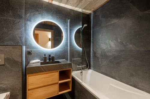 Modern bathroom with LED-lit round mirror, dark tile walls, wooden vanity, and white bathtub in Altitudia Chalet Magy 3