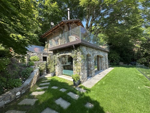 Luxury stone spa pavilion with glass terrace overlooking Lake Maggiore, surrounded by manicured gardens and tall trees at