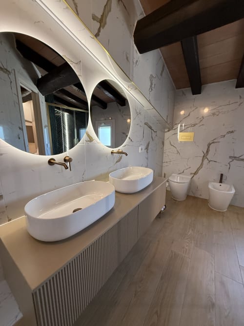 Suite bagno moderna a Borgo Albador con doppi lavabi bianchi, specchio rotondo, pareti in marmo, pavimento in legno e ottone