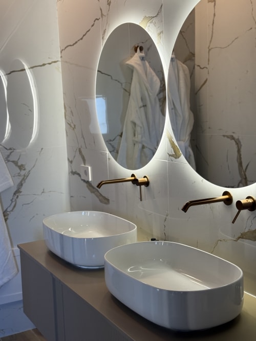 Bagno di lusso moderno nella Suite Borgo Albador a Torpè con doppi lavabi bianchi, specchio ovale illuminato e pareti in marmo
