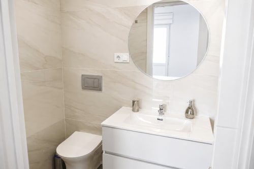 Nowoczesna łazienka z białą szafką, okrągłym lustrem i beżowymi płytkami na ścianach w apartamencie Alquileres Cantos de Sirena