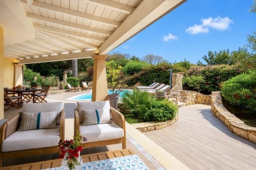 Luxury villa terrace in Sardinia with wooden seating, pergola, pool view, and Mediterranean gardens under clear blue sky