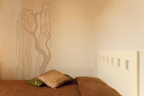 Camera singola presso Agorà Calamandrana con cuscino rigato, letto in legno e murale artistico in tonalità terra calde.