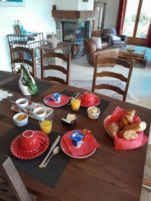 Table de petit-déjeuner au Mare e Monti Vico B&B à Vico avec assiettes rouges, croissants, jus d'orange et salon avec cheminée en arrière-plan.