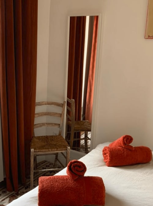 Chambre douillette avec serviettes rouges roulées sur literie blanche, fauteuil en osier, miroir et rideaux rouille au Mas des Ecoliers
