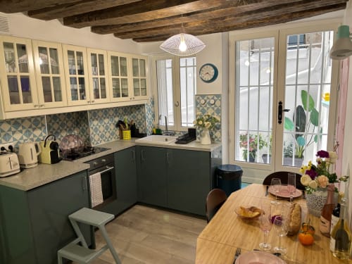 Cocina de apartamento parisino moderno con armarios verdes, salpicadero geométrico, vigas vistas, mesa de comedor y vistas al jardín