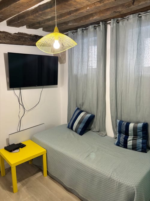 Dormitorio de apartamento parisino moderno con TV de pantalla plana, lámpara colgante amarilla, almohadas a rayas y cortinas grises en Aldaro