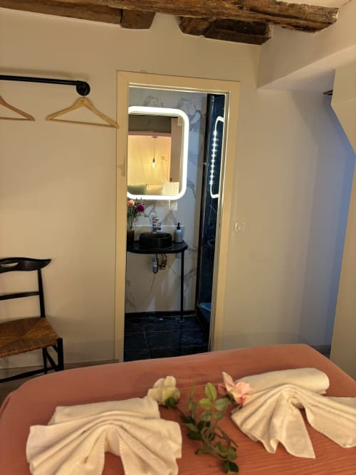 Acogedora habitación de apartamento parisino con puerta de baño, espejo iluminado, vigas de madera y toallas dobladas en la cama