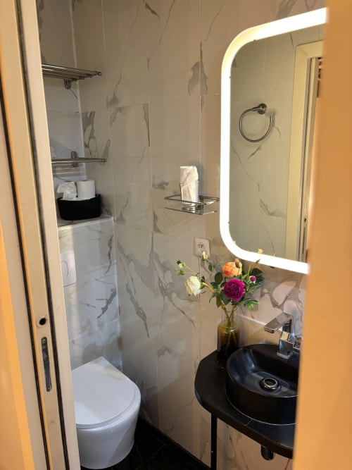 Baño moderno en apartamento parisino con lavabo negro, espejo iluminado, azulejos de mármol, flores frescas y ducha abierta