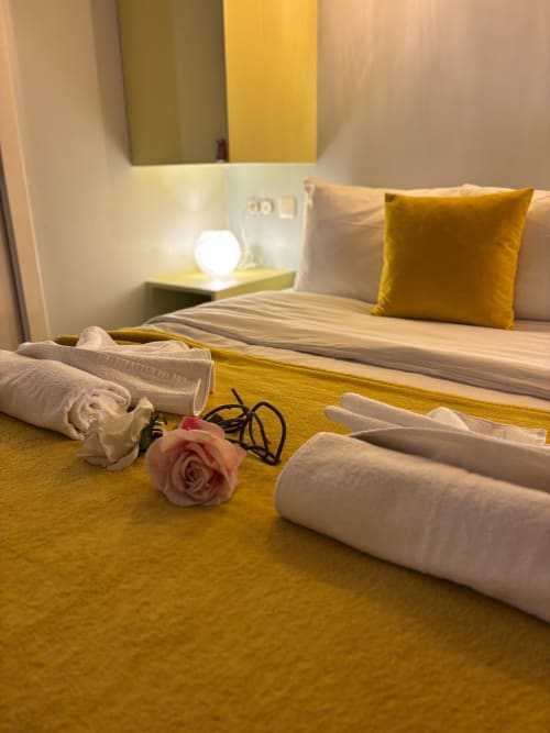Dormitorio de lujo en ALDARO Paris con camino de mesa amarillo, toallas blancas enrolladas, rosa rosa y almohada dorada sobre ropa de cama a rayas