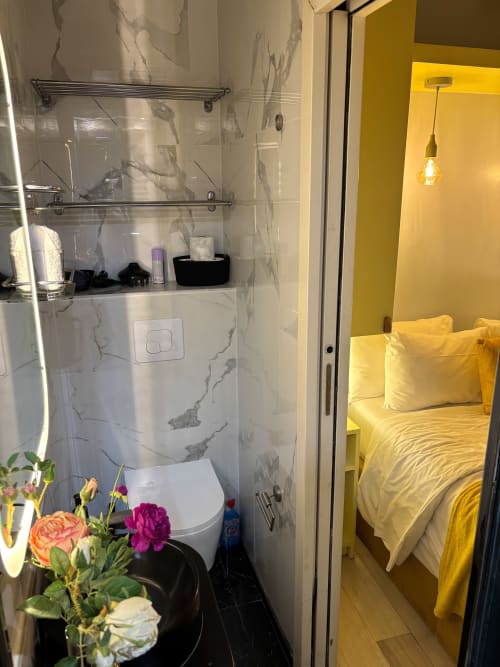 Baño moderno en apartamento parisino con baldosas de mármol, inodoro, cabina de ducha de vidrio y dormitorio amarillo visible