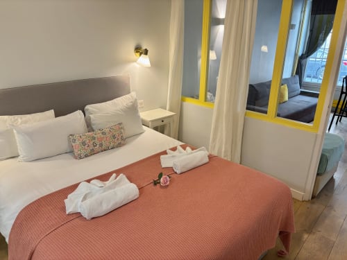 Dormitorio moderno en apartamento parisino con almohadas blancas, colcha coral, toallas enrolladas y puertas amarillas hacia la sala de estar