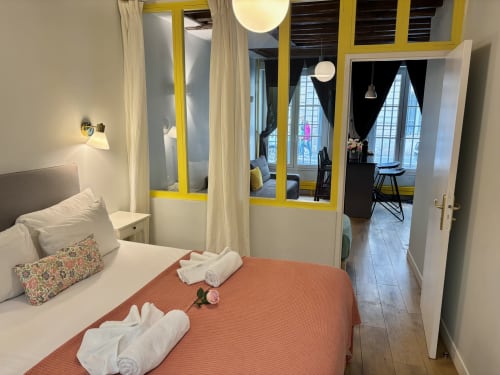 Dormitorio de apartamento parisino moderno con cama naranja, tabique de vidrio con marco amarillo, almohadas blancas y acceso a la sala de estar