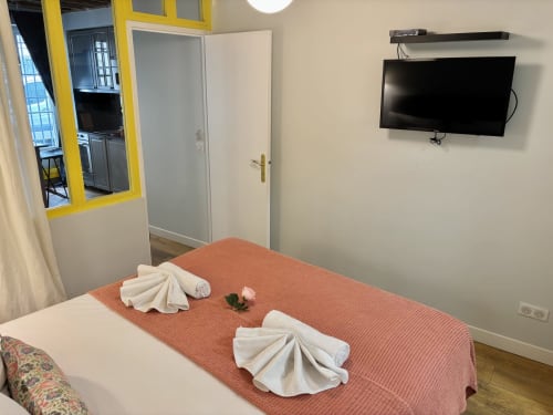 Dormitorio de apartamento parisino moderno con colcha naranja, toallas blancas, TV montada en pared y ventana con marco amarillo brillante