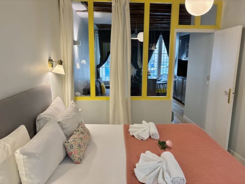 Dormitorio moderno en apartamento Aldaro Paris con colcha naranja, sábanas blancas, puerta acristalada marco amarillo y vista a la sala