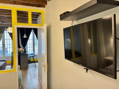 Interior moderno de apartamento parisino con TV montada en pared, puerta de marco amarillo hacia comedor, estante flotante gris