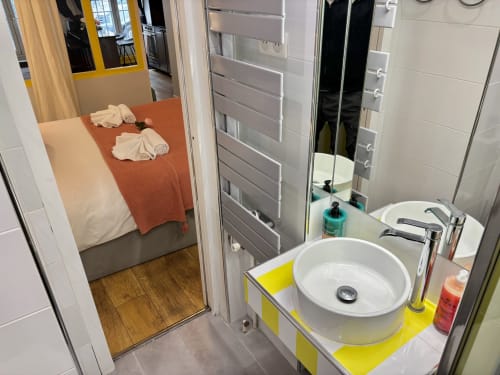 Baño moderno con lavabo redondo blanco sobre tocador amarillo, adyacente a dormitorio con manta naranja, apartamento Odéon París