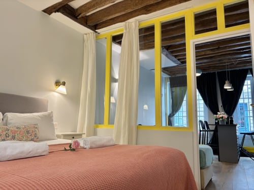 Dormitorio luminoso en apartamento parisino con ventanas marco amarillo, cortinas color crema, colcha coral y vigas de madera vistas
