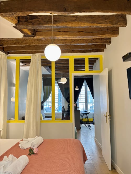 Dormitorio de apartamento parisino moderno con vigas de madera vistas, puerta de cristal con marco amarillo, colcha naranja e iluminación contemporánea