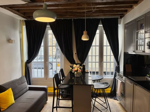 Comedor de apartamento parisino moderno con sillas negras, sofá gris, grandes ventanales, vigas vistas e iluminación colgante