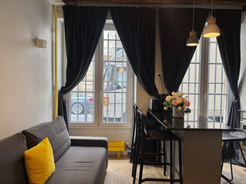 Moderno salón de apartamento parisino con sofá gris, cojín amarillo, cortinas negras, zona de comedor y arreglo floral
