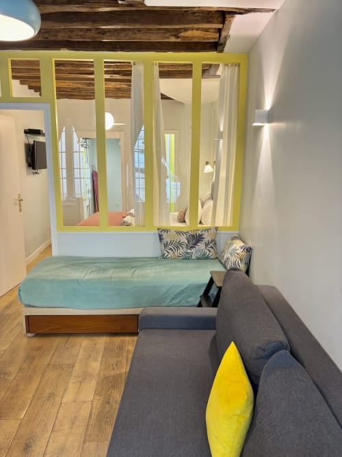 Dormitorio de apartamento parisino moderno con cama queen, sofá gris, acentos amarillo-verde, vigas expuestas y pared divisoria de vidrio