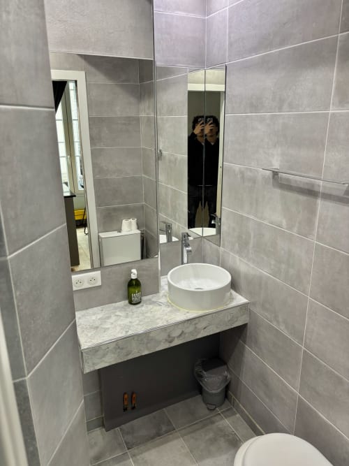 Baño moderno con lavabo blanco redondo, encimera de mármol, azulejos grises, espejo e inodoro en apartamento parisino