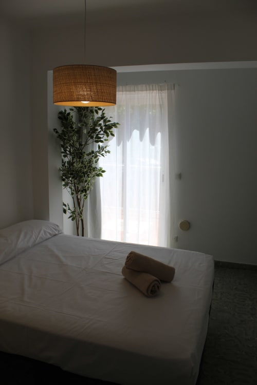 Luminoso dormitorio moderno en Villa Palmer hostel de Paguera con cama blanca, lámpara colgante de madera y acceso a balcón con cortinas semitransparentes