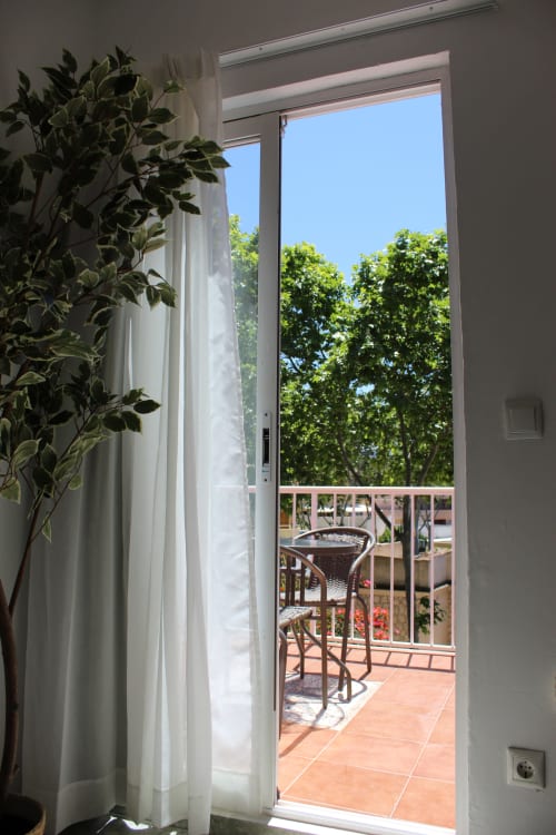 Vista radiante del balcón de la habitación Villa Palmer en Paguera con planta en maceta, cortinas translúcidas, silla de mimbre y exuberante vegetación