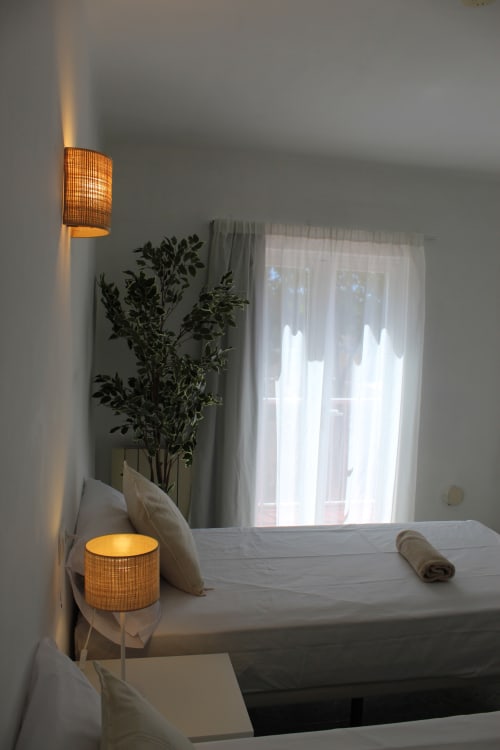 Dormitorio luminoso en Villa Palmer hostel de Paguera con ropa de cama blanca, iluminación ambiental, planta en maceta y cortinas translúcidas