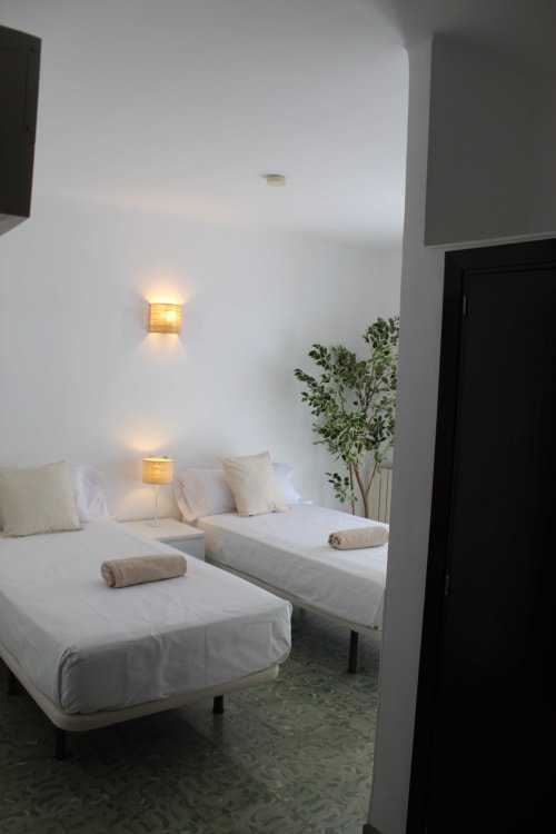 Dos camas gemelas con sábanas blancas en la moderna habitación del hostel Villa Palmer en Paguera, con iluminación ambiental y decoración con plantas