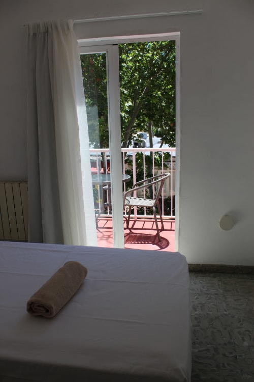 Dormitorio con cama blanca y toalla enrollada que abre a balcón soleado con silla y vistas al mar en Villa Palmer, Paguera