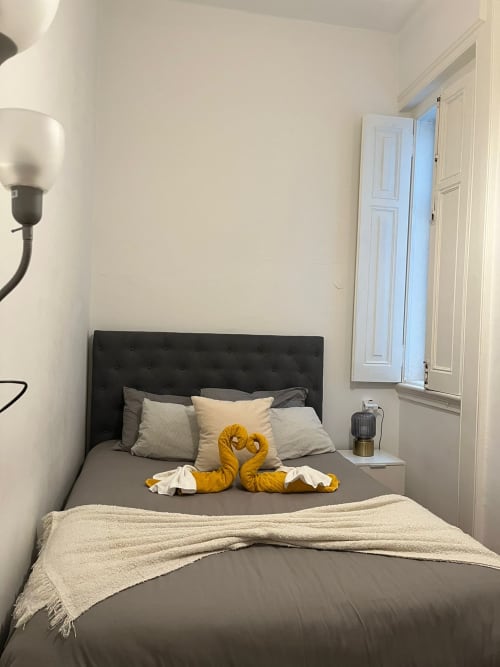 Quarto aconchegante de hostel em Lisboa com cabeceira preta, roupa de cama cinza, cisnes decorativos amarelos e manta creme
