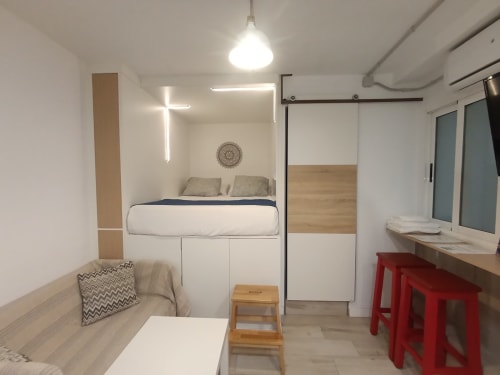 Dormitorio moderno y compacto con cama elevada, sofá, escritorio con taburetes rojos y cruz de neón en El Rincón de Sevilla