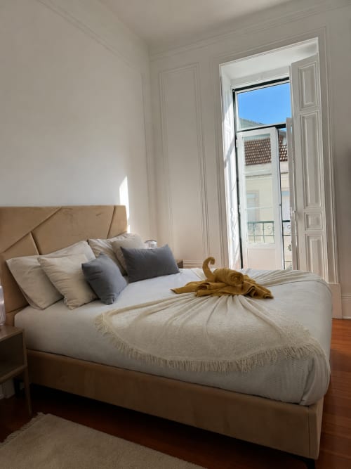 Quarto luminoso no hostel Grupo Lisboa Alojamentos com cama estofada bege, roupa de cama branca e portas abertas com vista para Lisboa