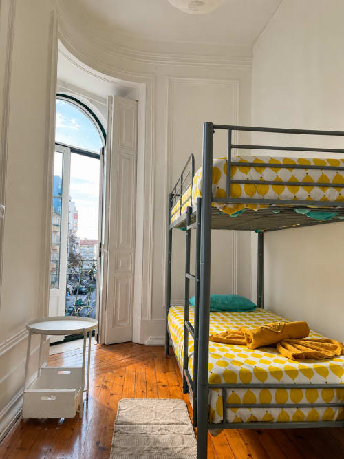 Quarto de dormitório luminoso em Lisboa com camas beliche pretas, roupa de cama com padrão amarelo, assoalho de madeira e janelas em arco