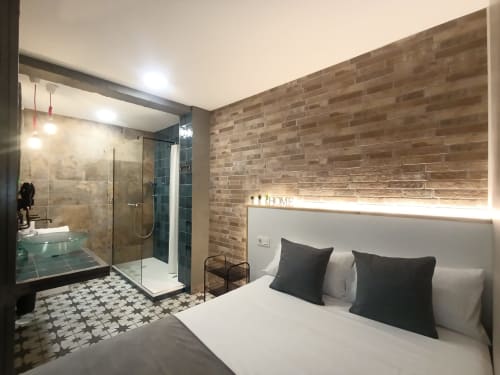 Moderno dormitorio con baño privado en El Rincón de Sevilla. Cama con almohadas negras, pared de ladrillo, cristal.